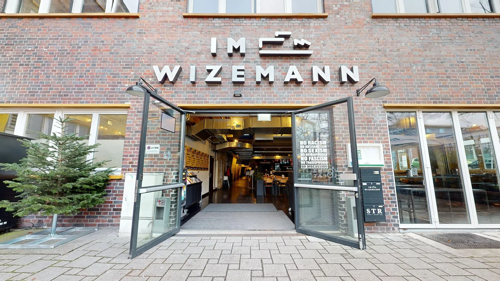 Eingang von Im Wizemann in Stuttgart, Backsteinfassade mit Leuchtschriftzug „IM WIZEMANN“ über den offenen Glastüren, Blick in den Gastrobereich